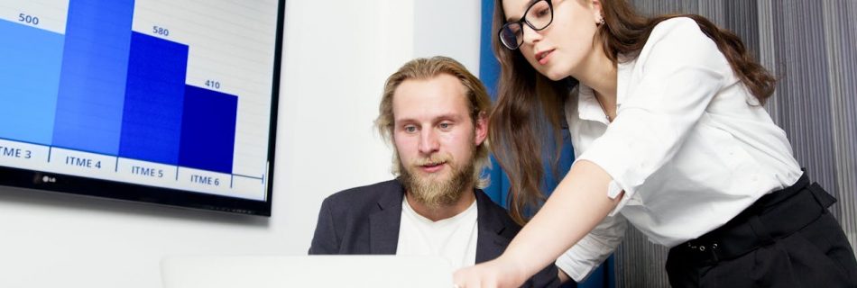 Two colleagues analyzing business data on a laptop with a presentation screen in the background.